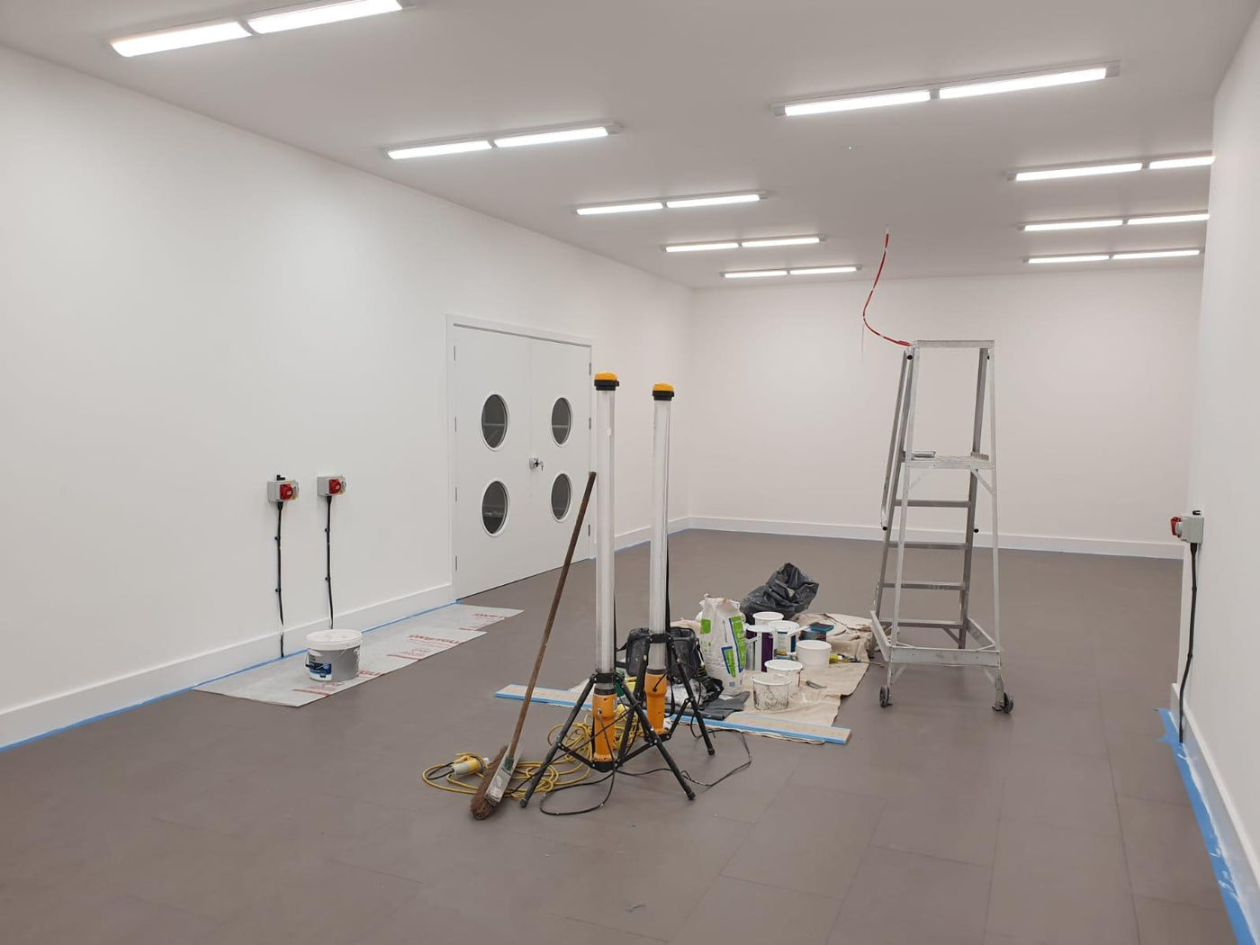 Empty room with white walls, gray floor, ladder, and painting supplies.